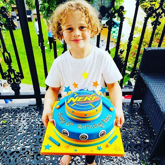 a curly haired boy holds a bright yellow and blue cake which says nerf on it and there are star shaped ornaments stuck into the top of it