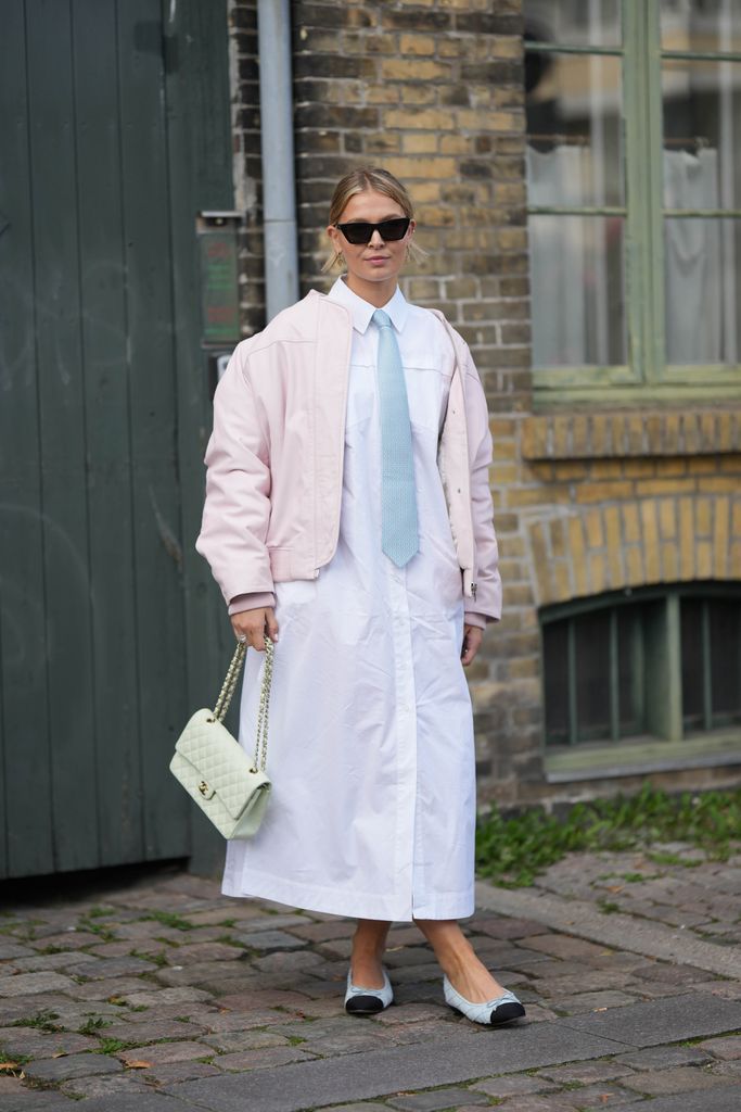 A guest wears black sunglasses, a pale pink oversized bomber coat, a pale blue tie, a white long slit shirt dress, a pale green matte leather handbag from Chanel, pale blue quilted leather and black toe cap ballerinas from Chanel , outside Remain, during the Copenhagen Fashion Week Spring/Summer 2024 on August 08, 2023 in Copenhagen, Denmark. (Photo by Edward Berthelot/Getty Images)
