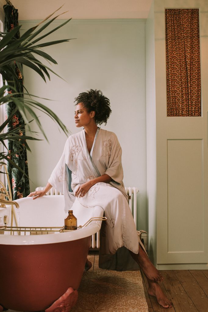 A woman perches on the side of an elegant red roll top bathtub, and waits for it to fill up. 