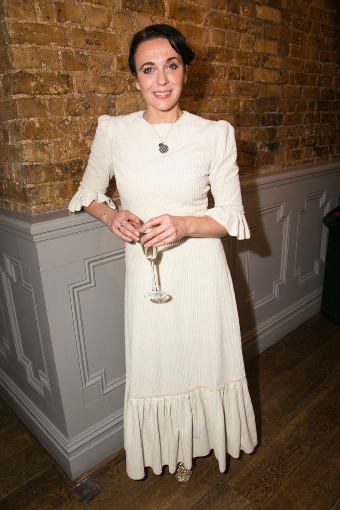 Amanda Abbington wearing a white dress against a brick wall background