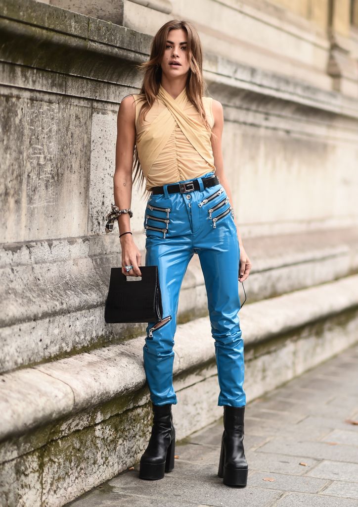 A fashion week guest wears a pair of blue latex skinny trousers with zip detail, black platform boots and a pale beige coloured halterneck top
