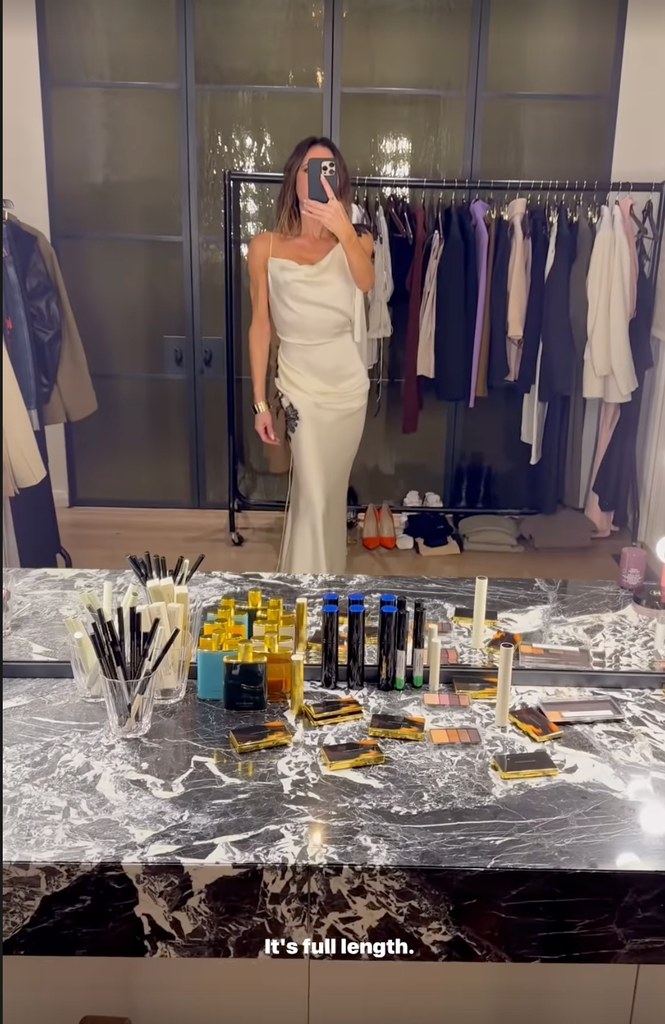 Victoria Beckham stands in her dressing room wearing a full length cream slip dress with embroidery on the sides and shoulder