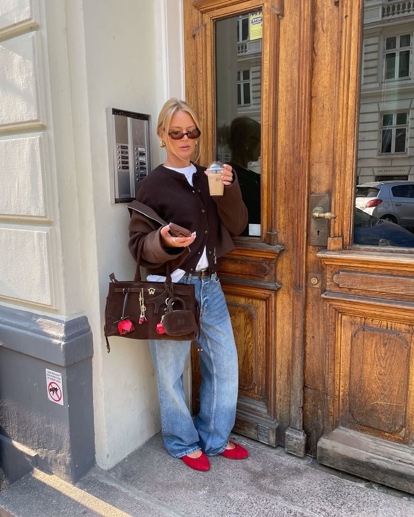 Danish style star Barbara Kristoffersen leans by a wooden door holding iced coffee, dressed in a chocolate cardigan, relaxed jeans, red ballet flats and a brown suede bag with rose embellishments.