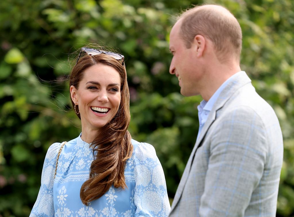 Princess Kate in a blue dress smiling while looking at her husband Prince William with green trees in the background