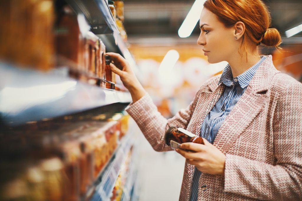 Young woman holding a jar of Sun-dried tomatoes in supermarket. She's searching for specific manufacturer.