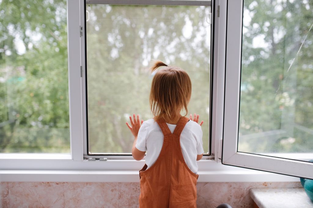 Toddler child looks out window and presses mosquito net with his hands. Child safety and protection