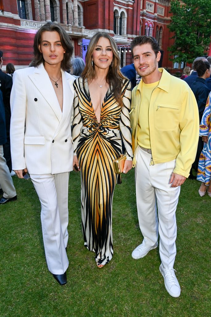 Damian and Elizabeth Hurley in a field alongside Gregg Sulkin in a yellow and white outfit