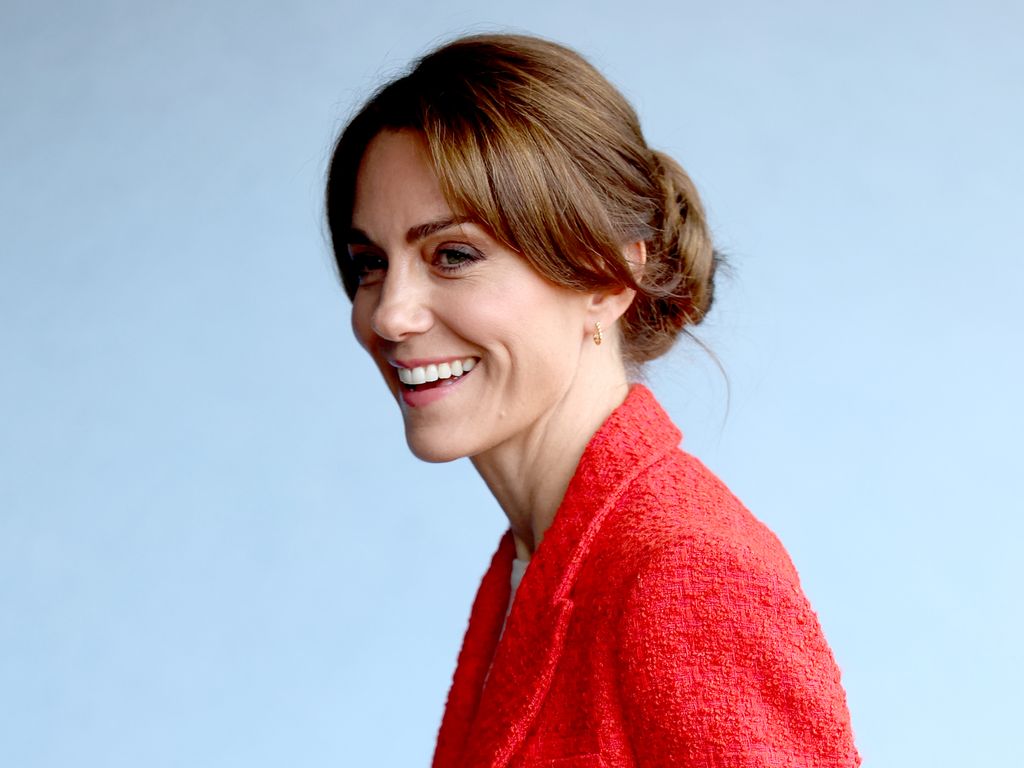 Princess Kate smiling wearing a red cardigan and her hair in a messy bun