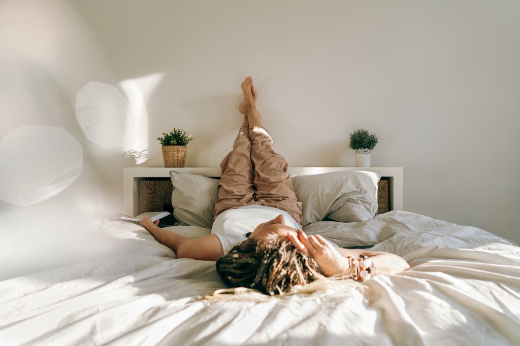 Woman in home clothes book lying on the bed reading. Work at home. Portrait of a dreadlocks braid woman legs up, inverted pose, swelling prevention varicose veins. High angle view