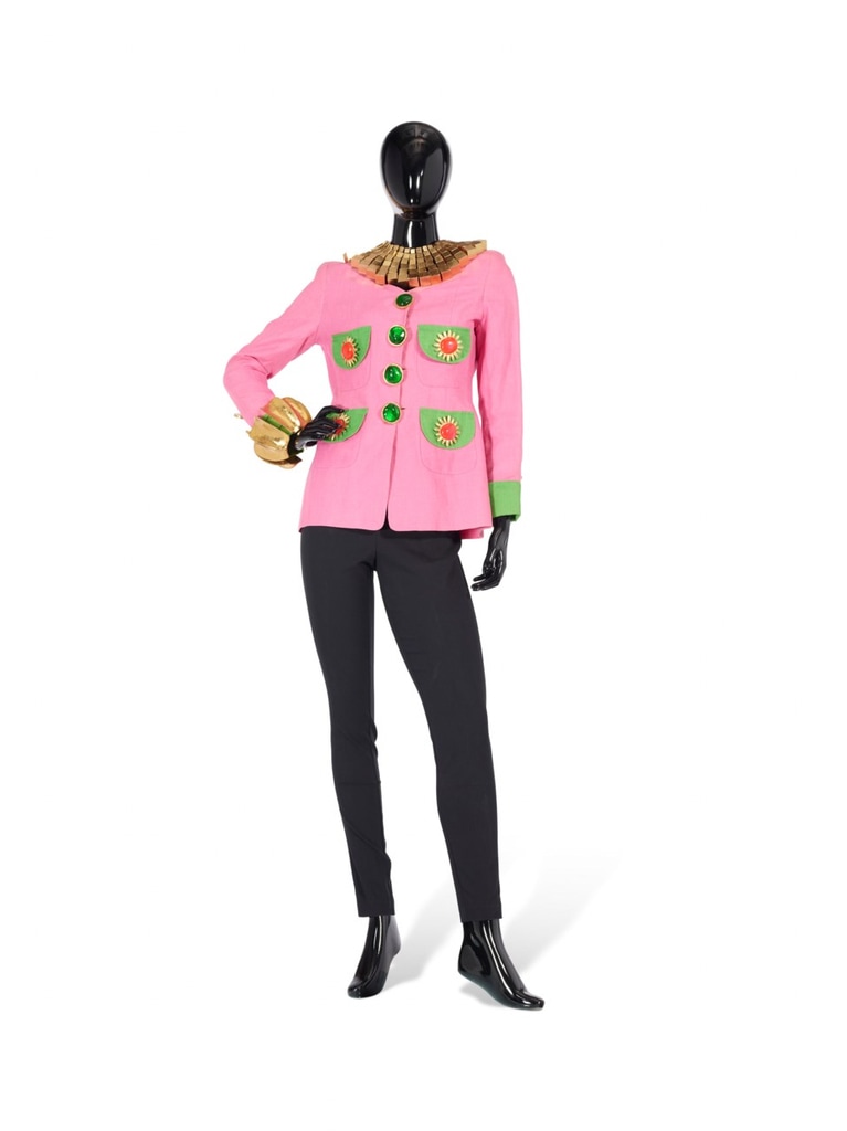 A pink jacket by Gemma Kahng (Circa 1990) with green pockets and floral buttons, creating a playful contrast. The jacket is paired with black pants, and the look is finished with a chunky, gold collar necklace by Monies, adding a bold touch to the playful ensemble. This joyful piece was part of Iris Apfel's wardrobe.