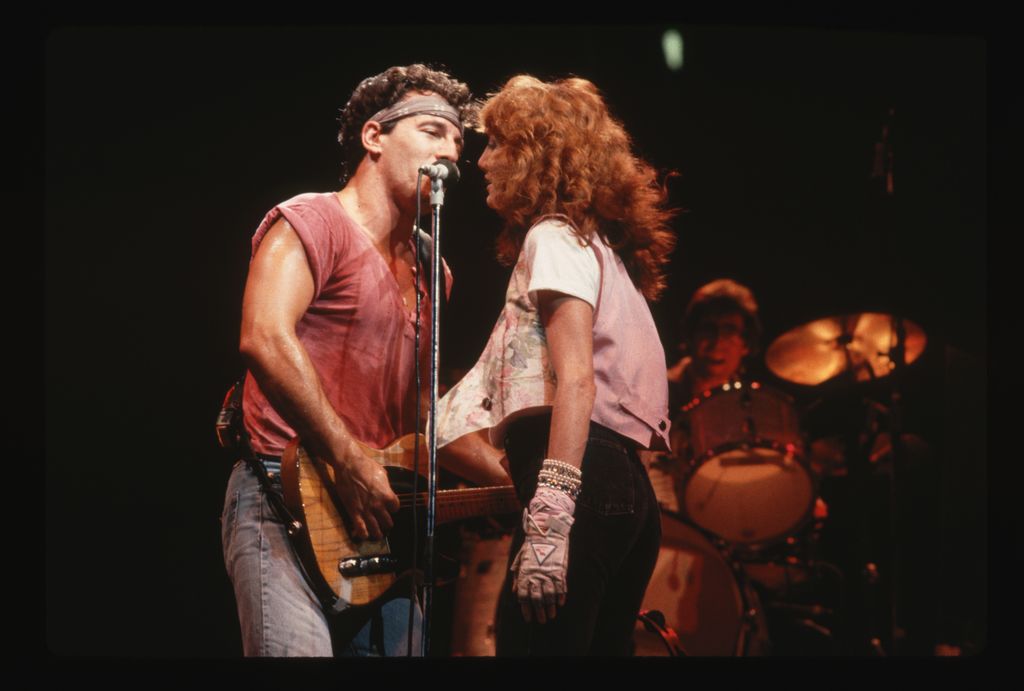 Bruce Springsteen on stage in concert, singing. He shares the microphone with back-up singer Patti Scialfa, whom he would go on to marry, 1984