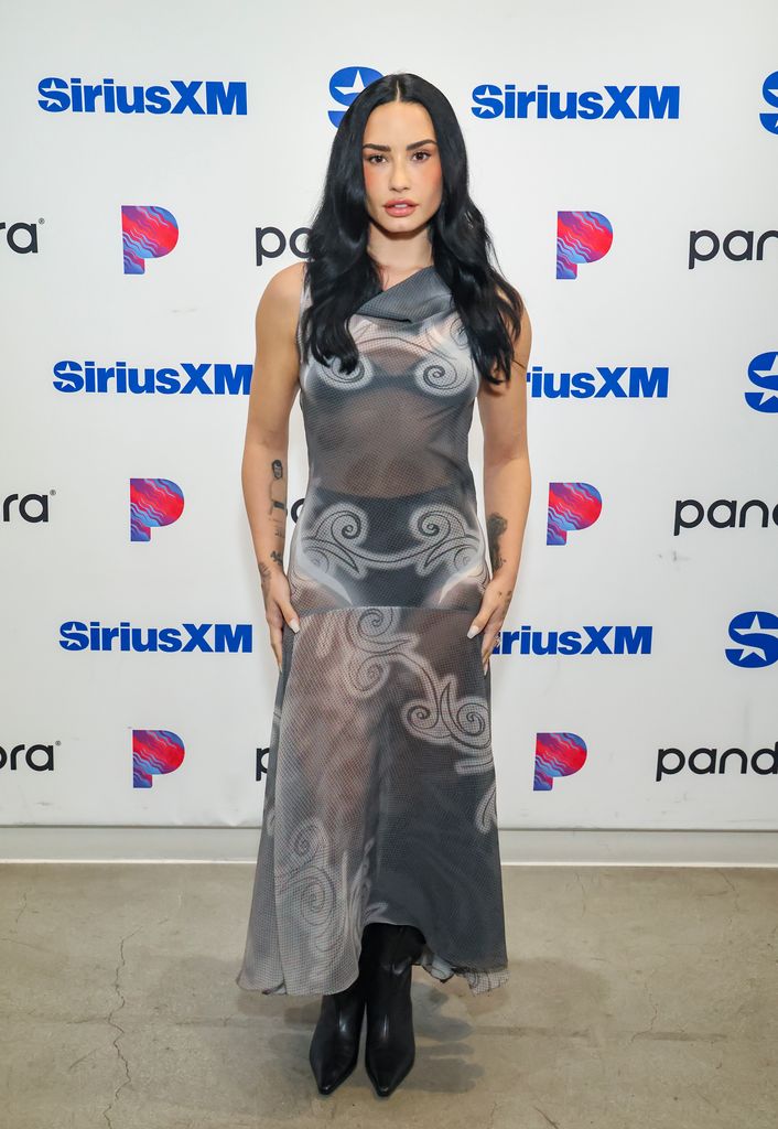 Demi Lovato poses in front of a backing board wearing a one shoulder grey sheer dress with celestial swirl detailing and black pointy boots.