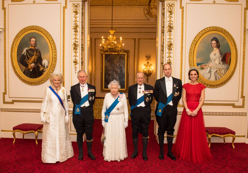 Kate em vestido vermelho ao lado da falecida Rainha, falecido Príncipe Philip, Charles, Camilla, William