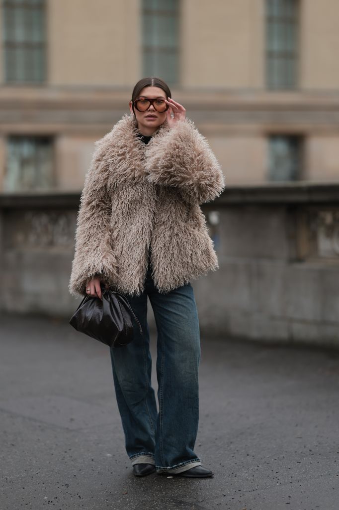 Sophia Geiss is seen wearing a large, textured, light brown or beige Source Unkown faux fur coat with a curly, shaggy appearance. The coat is paired with dark-wash, loose-fitting Closed denim jeans featuring a slight cuff at the hem. She is also wearing black, pointed-toe LeGer shoes and is holding a small black The Frankie Shop clutch purse in her right hand. She is wearing large, square-framed Jimmy Fairly sunglasses with a brown tint. Her hair is pulled back, and small hoop earrings are visible. The overall color palette is muted, combining the beige of the coat, the blue of the jeans, black accessories on December 10, 2025 in Berlin, Germany. (Photo by Jeremy Moeller/Getty Images)