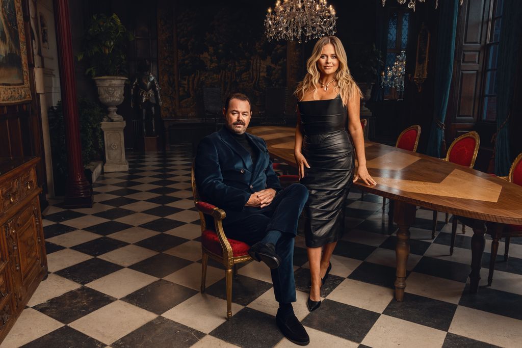A man in a suit sits on a chair and a woman stands next to him wearing a black dress