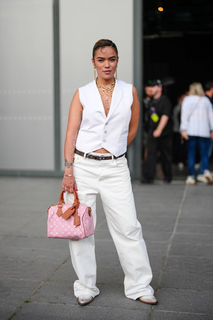 Karol G wears a white textured sleeveless vest with a deep V-neck and button fastening. Layered gold necklaces and large hoop earrings are visible. Hair is styled in a sleek, side-parted look. The outfit includes white high-waisted trousers with a black belt featuring a silver buckle. A pink handbag with a monogram pattern and brown leather handles is carried. Footwear consists of white shoes with a pointed toe, outside Louis Vuitton, during Paris Fashion Week - Menswear Spring/Summer 2026, on June 24, 2025 in Paris, France (Photo by Edward Berthelot/Getty Images)