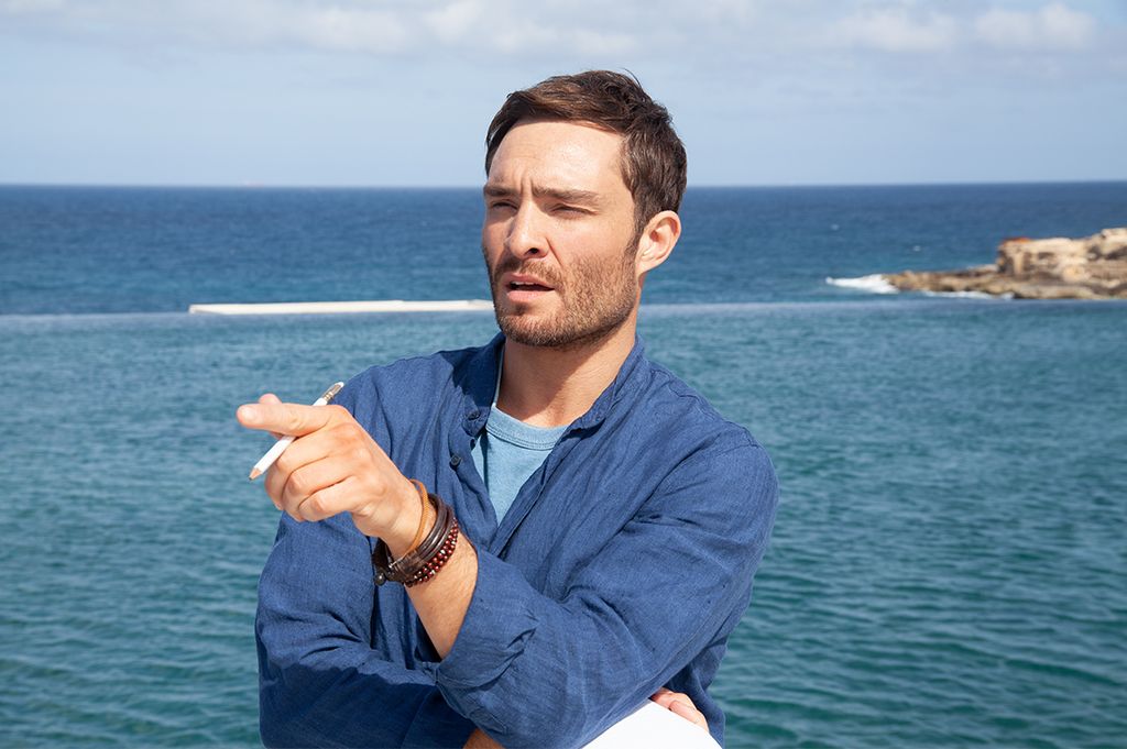 Ed Westwick posing against seaside backdrop in blue shirt