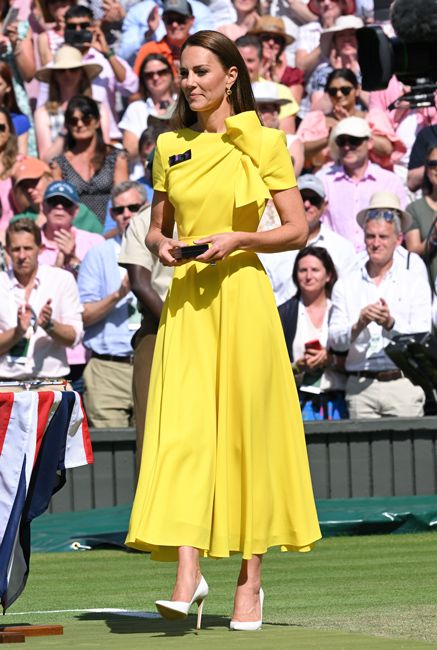 kate walks on a tennis court and stands out in a floaty mid length yellow dress with a large bow at the collar and her white high heeled shoes stand out