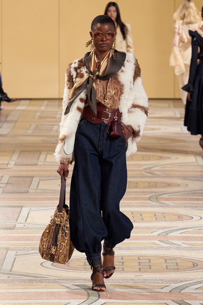 A model walks down the runway in a fluffy cow print coat, brown neck tie and denim trousers
