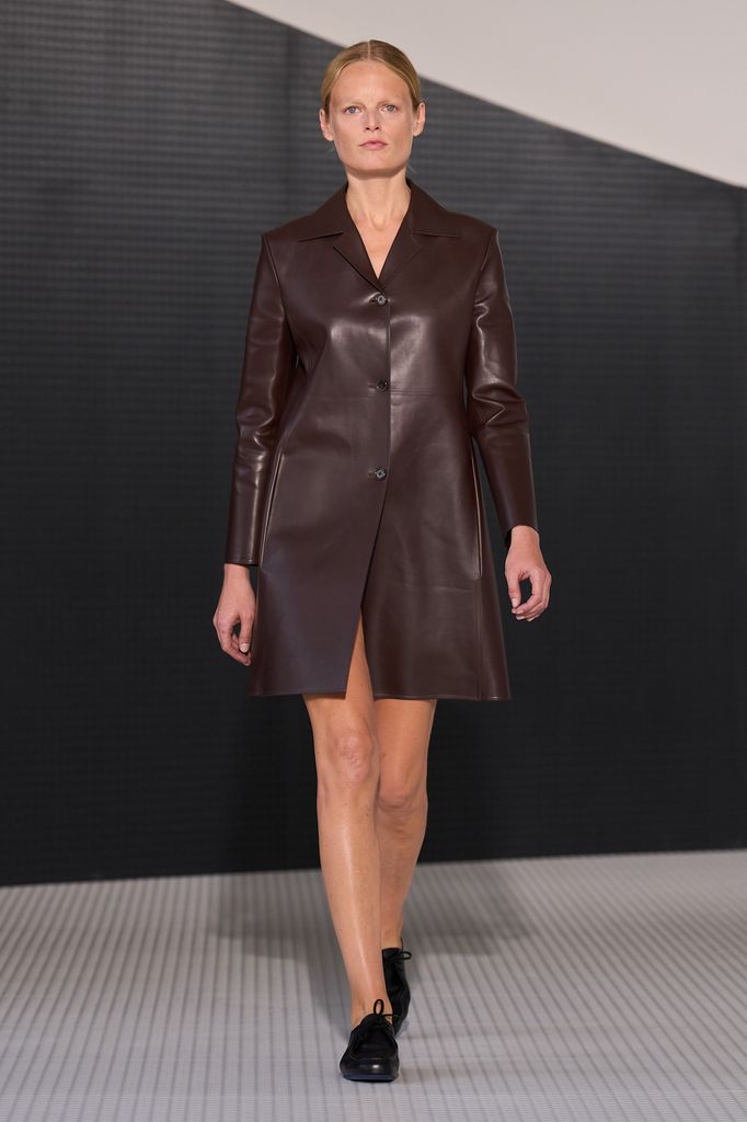 A model wears a dark brown long sleeved leather mini coat and black lace up shoes. 