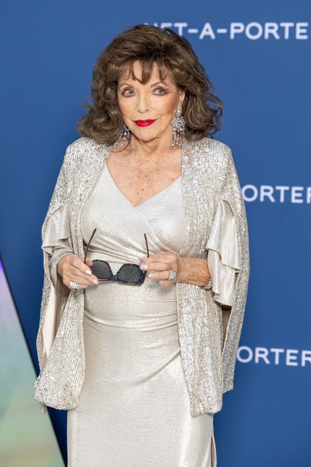 Joan Collins in a shimmering white outfit holding her sunglasses