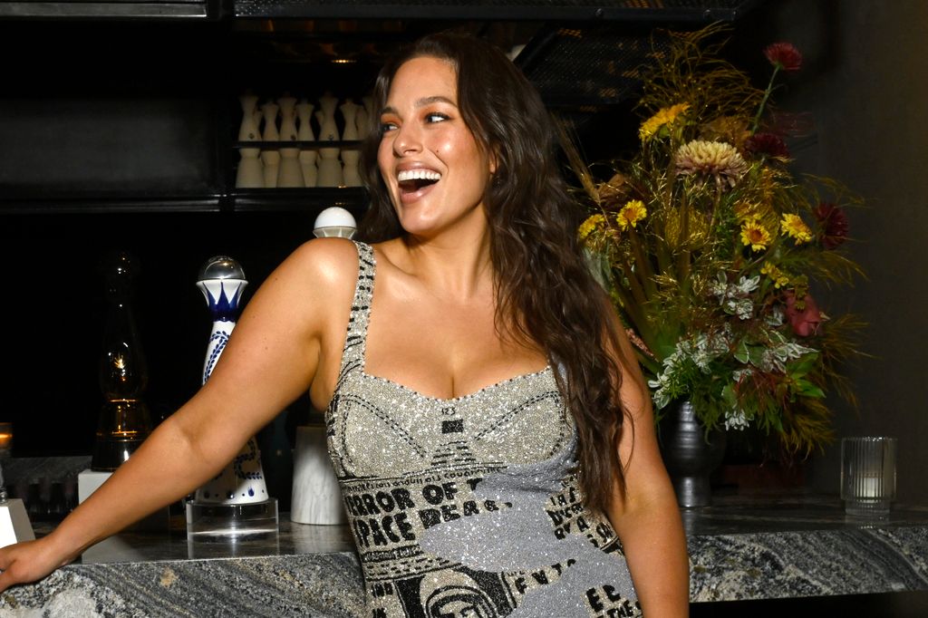 Ashley Graham poses on bar at Clase Azul event held at The Loft Brooklyn wearing sparkly silver mini dress