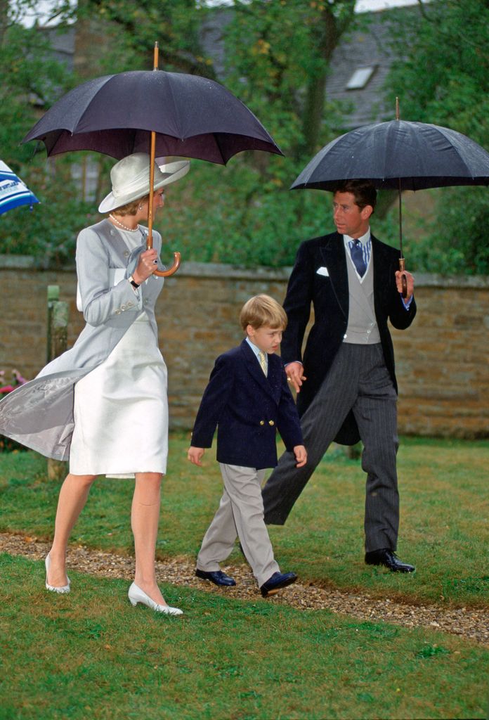 Princess Diana in a white dress and a grey coat under an umbrella with Charles and William