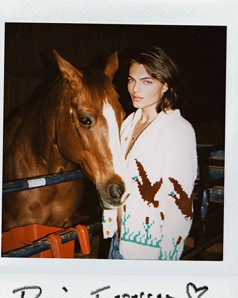 A photo of Damian Hurley wearing a cream cardigan and posing with a horse 