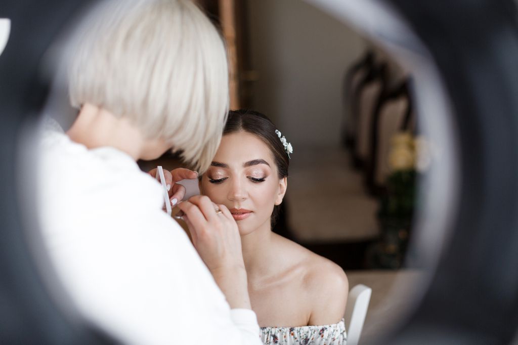 Beautiful young bride sits in the chair and looks down. Makeup artist puts eye shadows on the bride