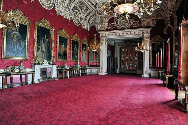 the a large room decorated with deep red carpet and matching wallpaper with small wooden tables pushed to the edges of the room below gold framed paintings
