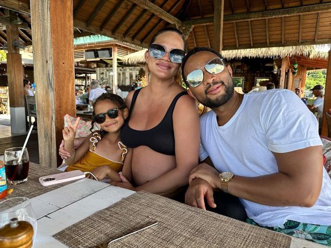 a heavily pregnant chrissy sits at a table under a cabana wearing a plunging black bikini top and sunglasses as a little girl and man sit beside her also wearing sunglasses as they smile at the camera
