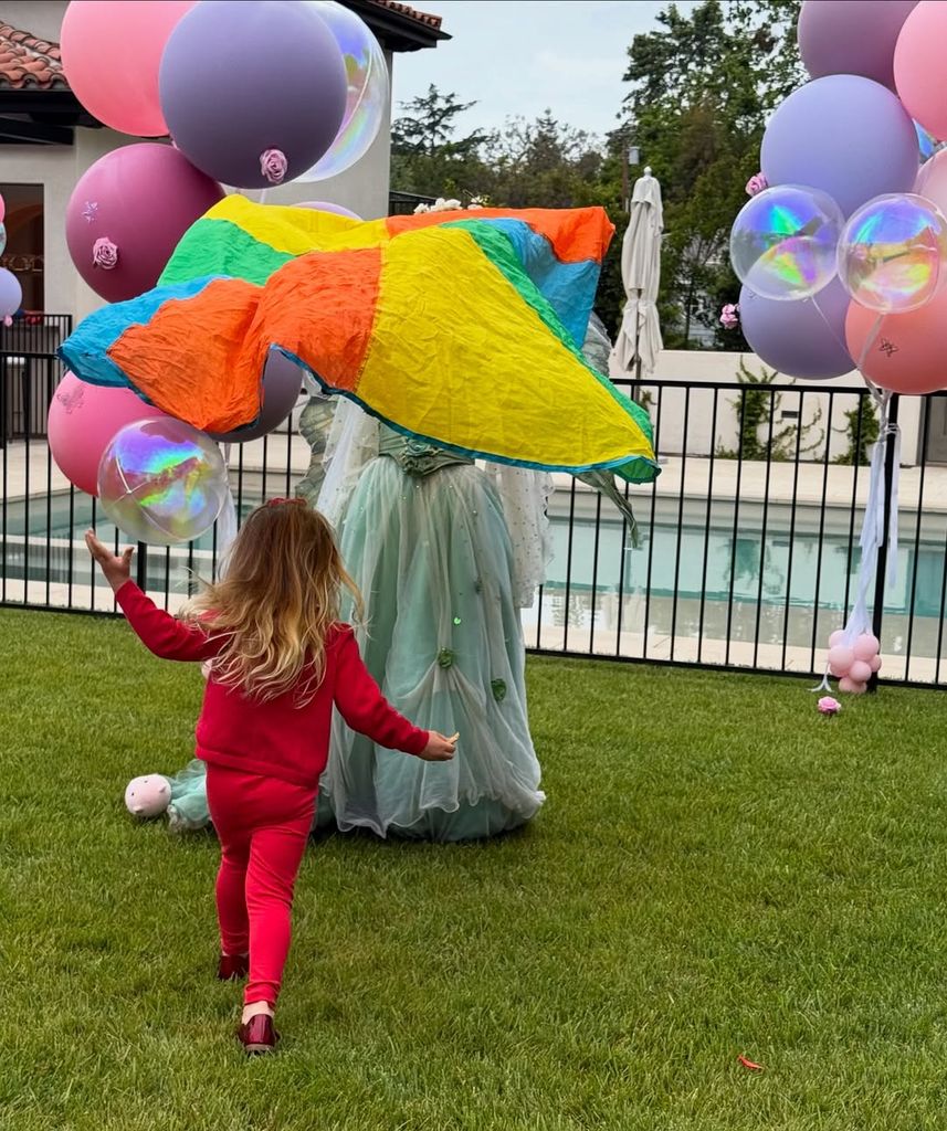 Young girl dressed in red running across the lawn towards princess figurine and colorful balloons