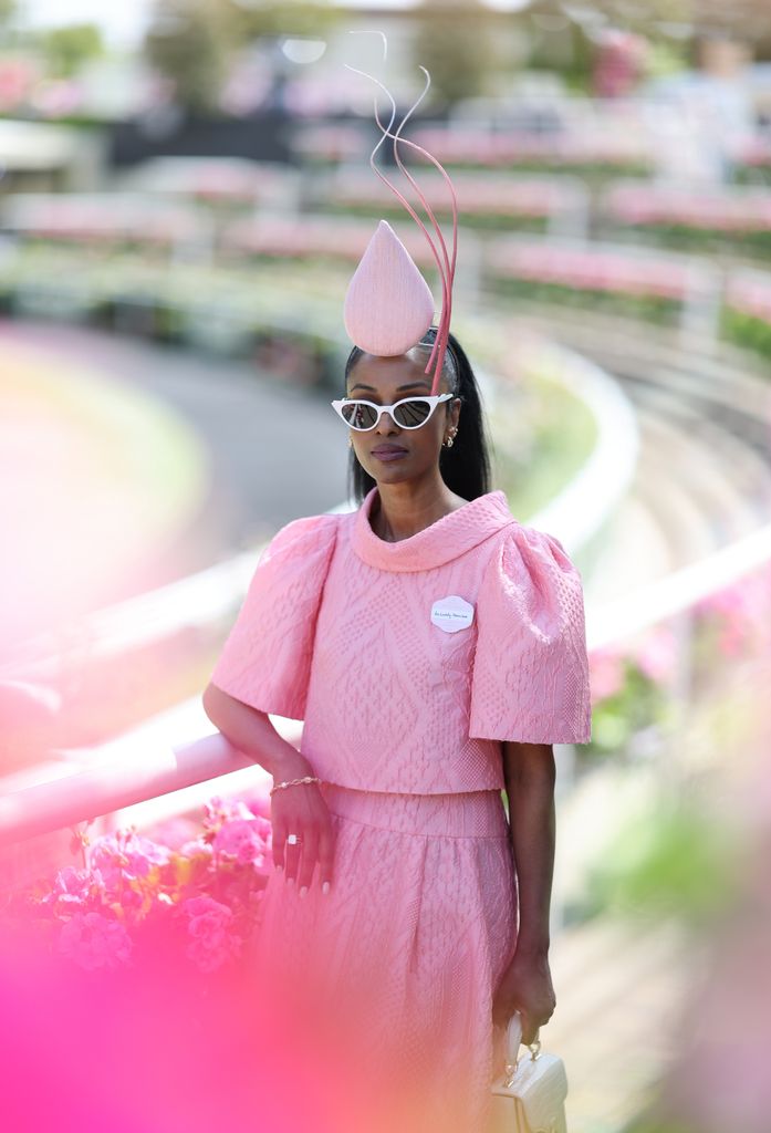 Lovely Amusan at Royal Ascot wears a textured pink dress with sculptural fascinator and white sunglasses in a sea of florals.