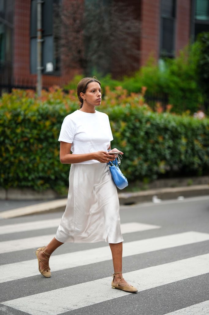 A guest wears a white t-shirt, a royal blue shiny leather puffy pockets handbag from Prada, white latte silk satin midi skirt, beige suede ballerinas , outside JW Anderson, during the Milan Fashion Week - Me