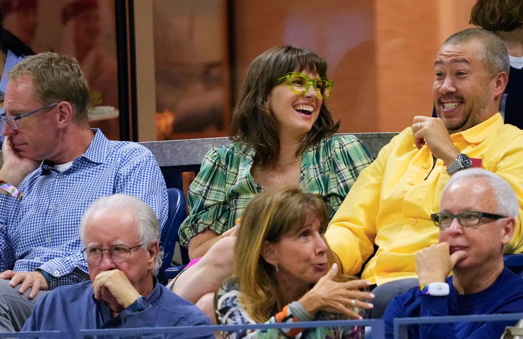Anna Chlumsky wears green glasses and laughs with her husband Shaun So at a sports game