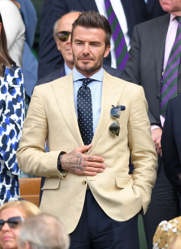 David Beckham stands in the crowd at Wimbledon wearing a pale tan blazer, blue shirt and dark blue polka dot tie. 
