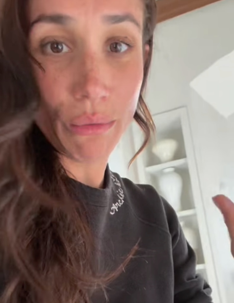 Meghan Markle is seen in a close-up selfie-style shot wearing a black sweatshirt with white cursive embroidery on the collar. Her hair cascades naturally, and she appears mid-conversation, her expression engaged and slightly amused. A shelf with white vases is in the softly lit background.