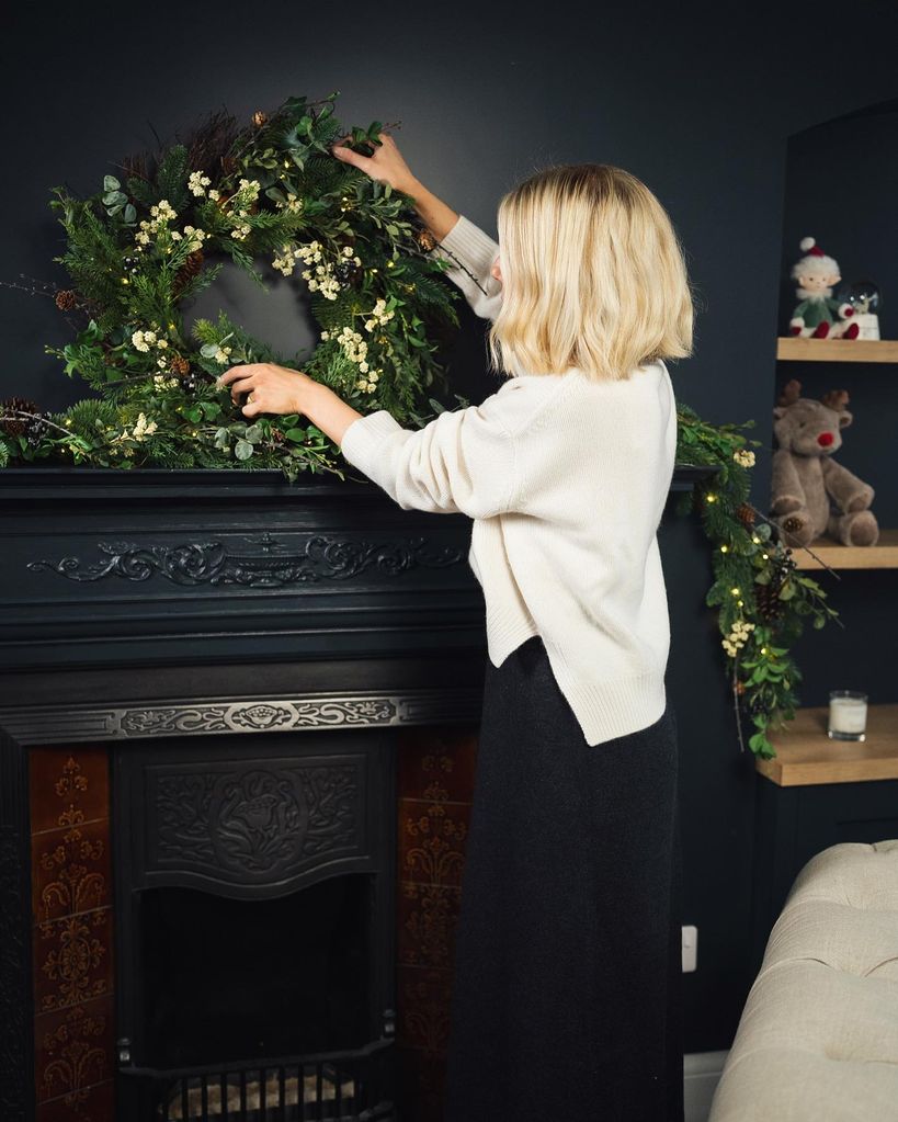 Mollie King wears a black satin skirt and white polo neck jumper as she arranges a wreath