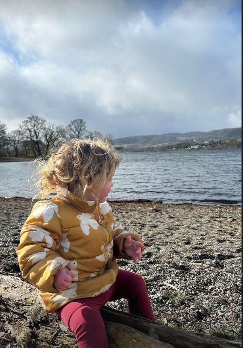 A blonde toddler sat on a log by a lake