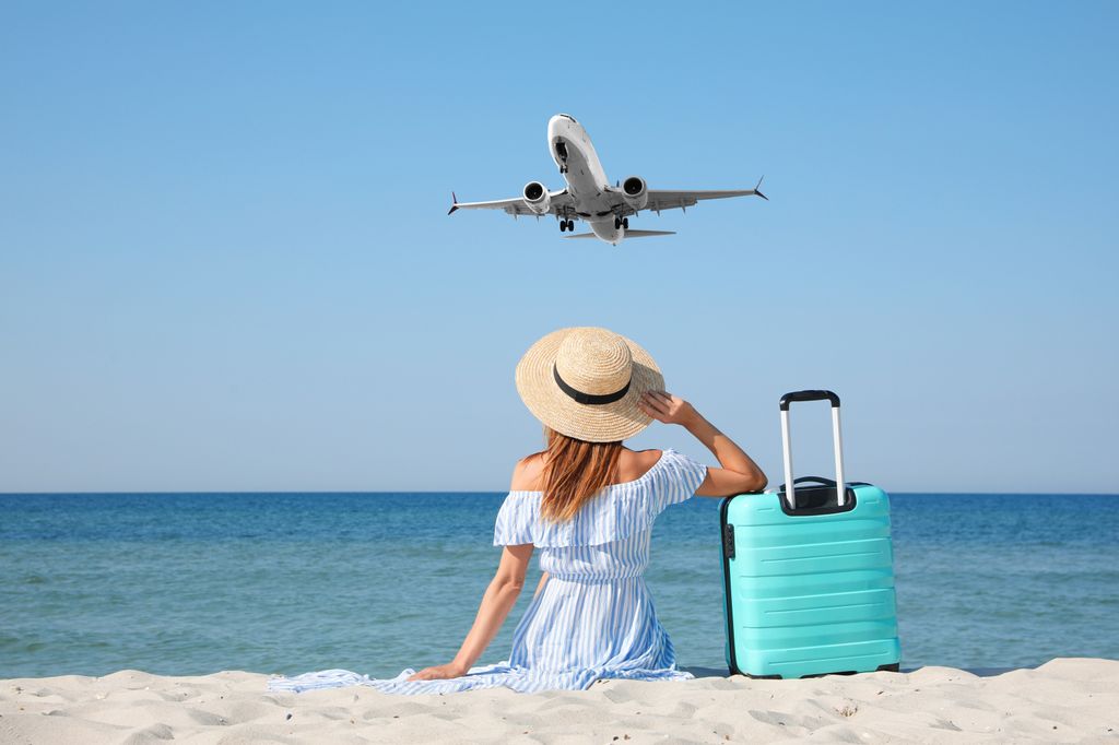 Woman with suitcase on sandy beach looking at airplane flying