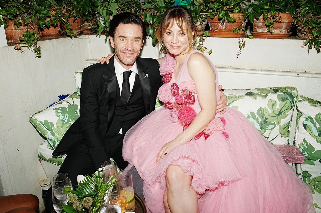 Tom Pelphrey in a suit sitting on a sofa with Kaley Cuoco in a pink dress