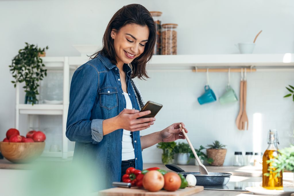 Shot of smiling beautiful woman using her mobile phone while cooking vegetables in the kitchen at home.