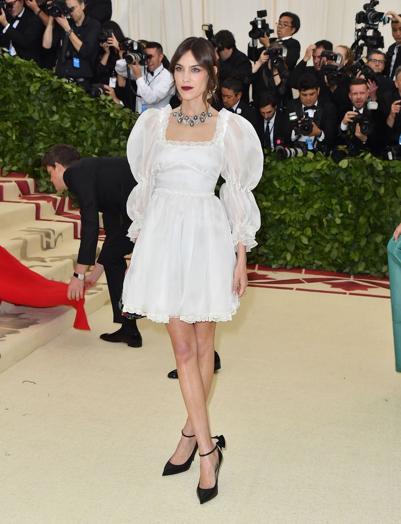 Alexa Chung wears a white dress with long puffy sleeves, a silver necklace and black heels with a bow at the back. Her hair is in a soft updo and her lipstick is deep and red. 