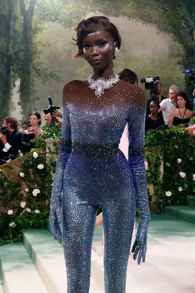 Anok Yai walks the 2024 Met Gala red carpet in a shimmering Swarovski crystal catsuit and oversized silver floral necklace with matching earrings, exuding futuristic glamour under garden-themed lighting