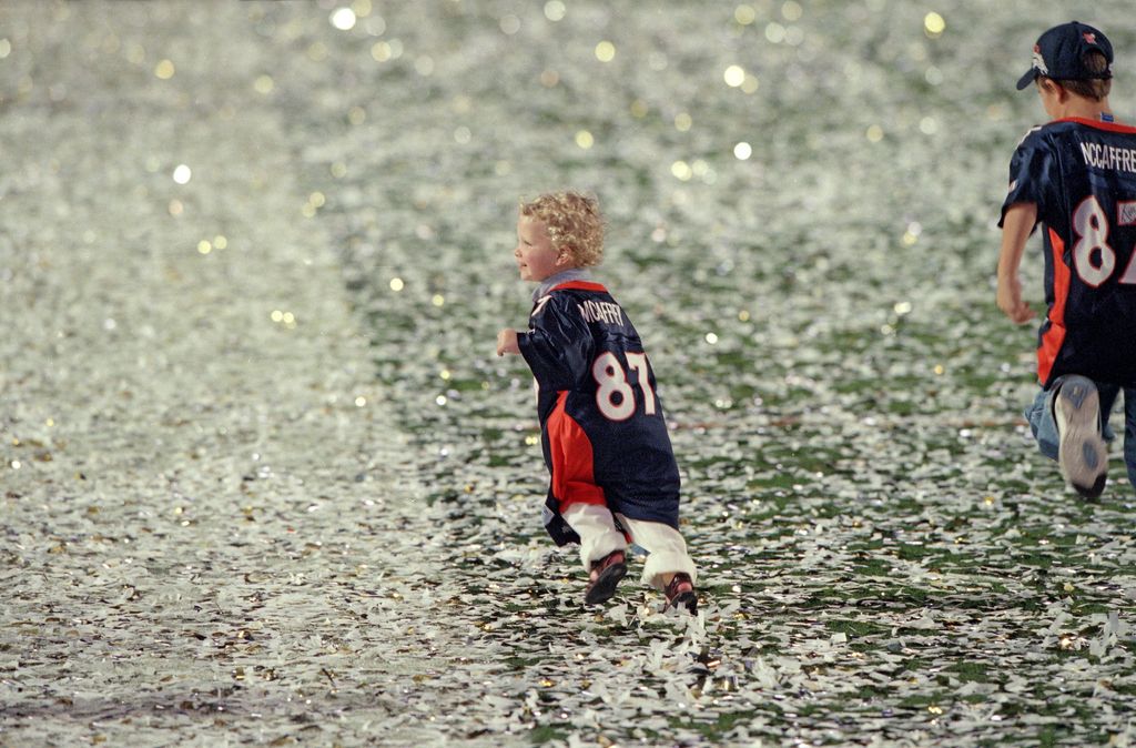 Christian McCaffrey (C) and Max McCaffrey (R), sons of Denver Broncos Ed McCaffrey, victorious and running on field in their father's jersey after winning game vs Atlanta Falcons at Super Bowl, 1/31/1999