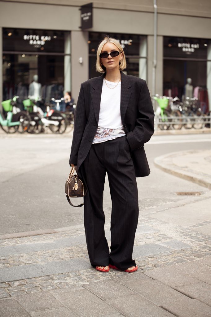 A guest wears white top, black pants, black blazer, white scarf over her hips and a brown bag and red flip flops outside Filippa K presentation during Copenhagen Fashion Week day four on August 07, 2025 in Copenhagen, Denmark.