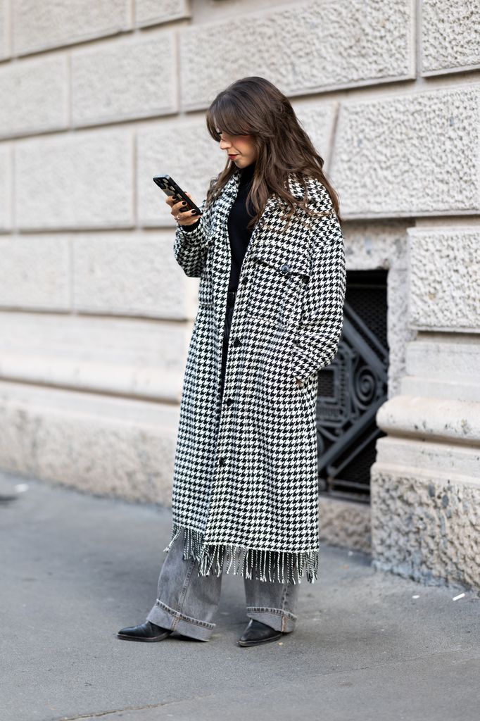A passerby wears a black-and-white houndstooth long coat with fringed hem over a black top and cuffed grey jeans, styled with black pointed boots and wavy hair with a fringe outside Pilot Room showroom on November 13, 2025 in Milan, Italy. (Photo by Valentina Frugiuele/Getty Images)