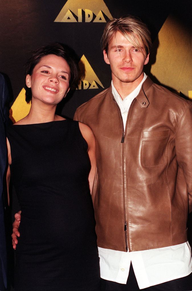 Victoria wearing a black dress smiling, David is wearing a brown leather jacket and white shirt with his arm around her