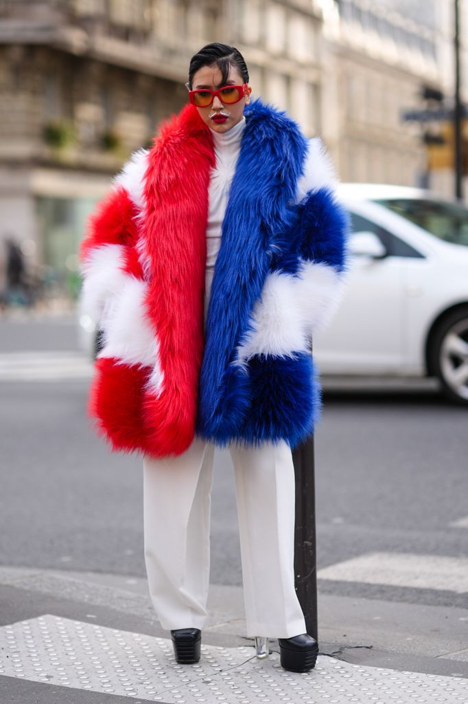 A guest wears red sunglasses, a white red and blue striped oversized fluffy faux fur coat , a white turtleneck top, wide leg white pants , platform black leather shoes, outside Casablanca, during the Womenswear Fall/Winter 2024/2025 as part of  Paris Fashion Week