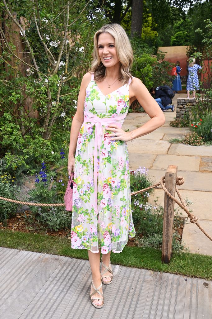 Charlotte Hawkins wears a floral pastel dress with pink ribbon belt and silver sandals poses in garden setting at Chelsea Flower Show 2025.